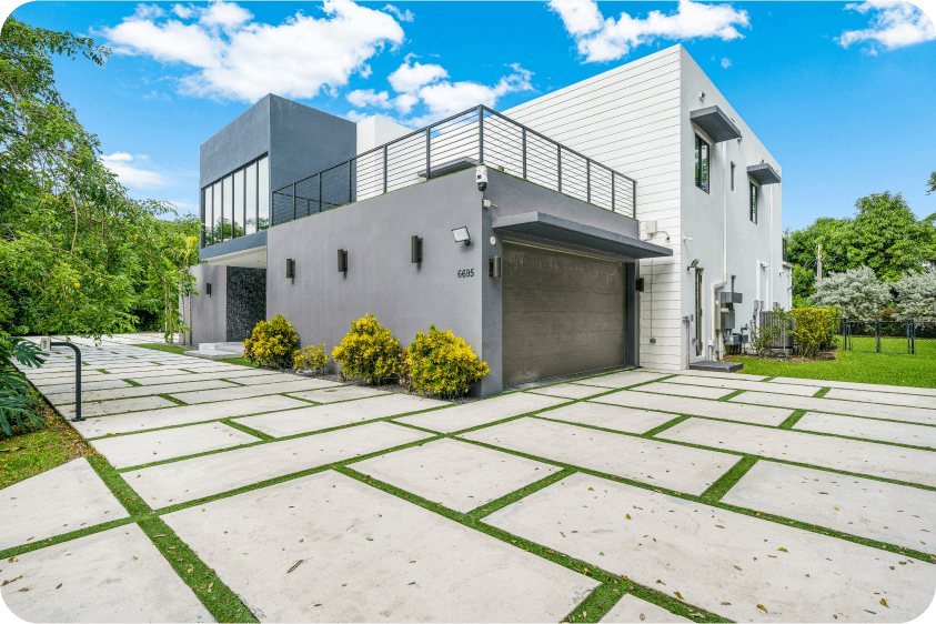 Modern side facade of luxury Pinecrest residence featuring balcony, garage door, and landscaped pavers with tropical greenery